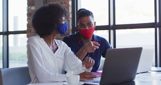 Colleagues wearing face masks working on project in a contemporary office with large windows. Ideal for illustrating remote work, contemporary workspaces, pandemic workplace safety, business collaboration, and health precautions in professional environments.