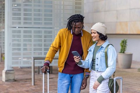 Multicultural young travelers checking phone at urban plaza with carry-on suitcases