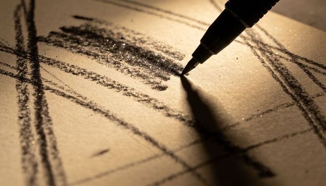 Fine-nib pen sketching charcoal strokes on textured paper casting dramatic shadow