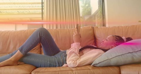 Young woman relaxing on leather sofa scrolling smartphone in sunlit modern living room