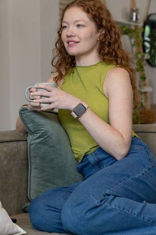 Relaxed redhead woman sipping coffee on sofa wearing lime sleeveless top and blue jeans