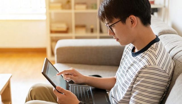 Young man using tablet and laptop on sofa at home studying, browsing and working remotely