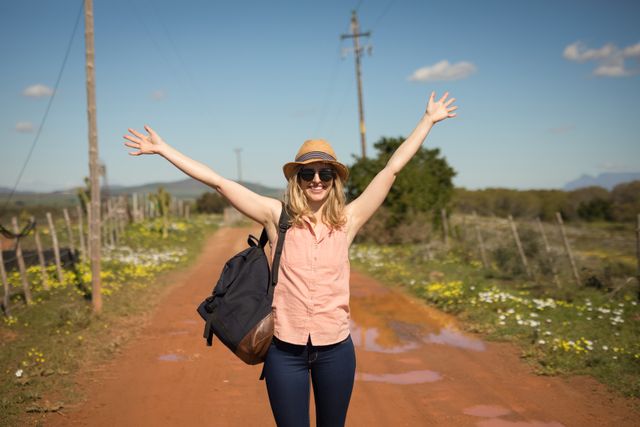 Woman enjoying a hike on a sunny day, walking along a rural path with arms raised in joy. Ideal for travel blogs, outdoor adventure promotions, and lifestyle content emphasizing freedom and happiness.