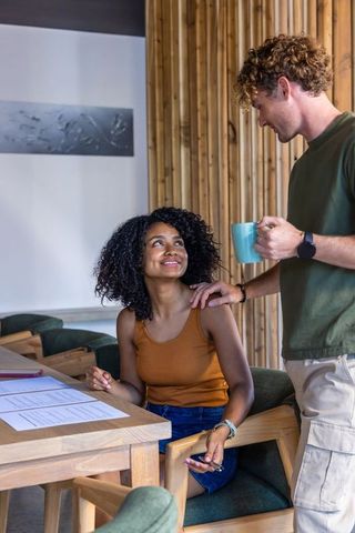Diverse coworkers smiling and chatting at wooden coworking table with coffee and documents