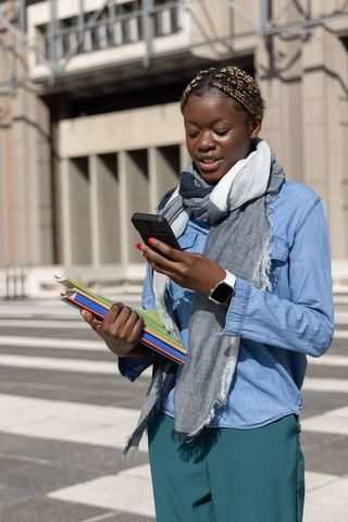 African american female student checking smartphone holding colorful notebooks on city crosswalk