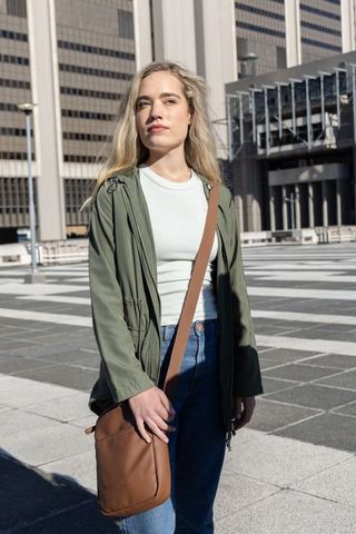 Woman standing in sunny urban plaza wearing olive jacket carrying brown crossbody bag