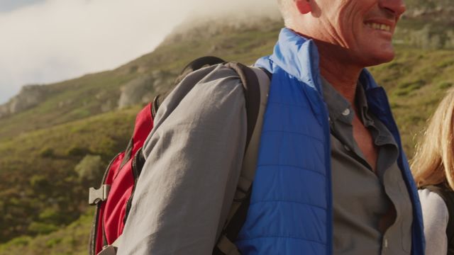 A senior couple enjoys hiking through a lush meadow with backpacks. The photo conveys a sense of adventure and appreciation for the outdoors. Ideal for use in promoting outdoor activities, senior lifestyle blogs, or travel content aimed at mature audiences.