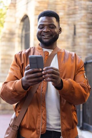 Smiling african man using smartphone near historic stone archway rust jacket portrait