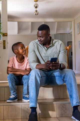 African american father and son sitting on steps smiling, using smartphone, family bonding