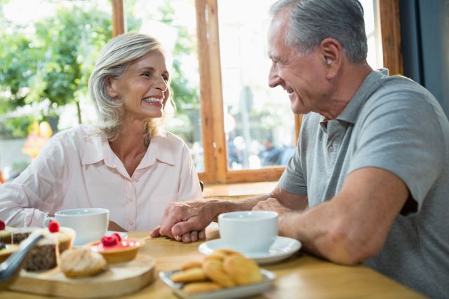 Senior couple sitting at a table in a cafe, enjoying coffee and pastries while smiling and interacting with each other. Ideal for use in advertisements for cafes, retirement communities, lifestyle blogs, and articles about senior living, relationships, and leisure activities.