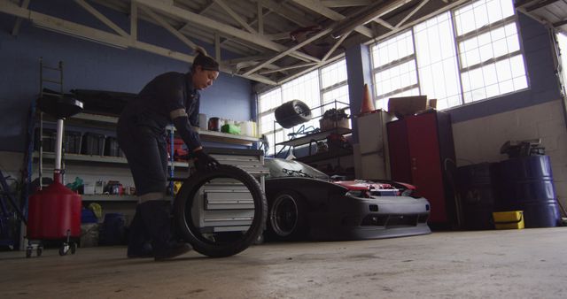 A professional mechanic dressed in work overalls is rolling a car tire in an auto repair garage. In the background, various tools and equipment are visible along with a partially repaired sports car with the hood open. This photo is ideal for use in automotive industry advertisements, repair service websites, technical training materials, and showcasing mechanic work environments.
