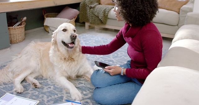 Afro-American woman wearing red turtleneck sitting on carpeted floor petting golden retriever in cozy, well-lit living room. Ideal for use in articles or ads promoting pet care, home comfort, relaxation, and bonds between pets and owners.