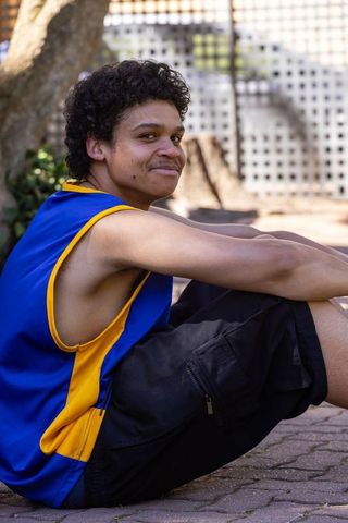 African-american teen sitting on courtyard pavers wearing blue yellow basketball jersey smiling