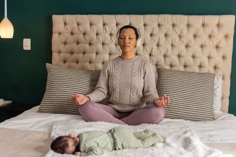 Mother meditating on bed while baby sleeps nearby, headphones, mindful parenting