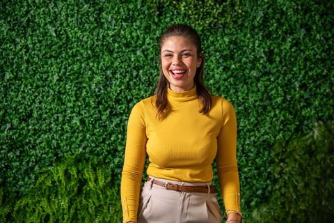Smiling woman standing by lush green hedge wearing mustard turtleneck and beige trousers
