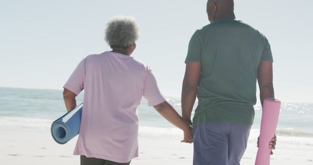Senior couple is holding hands while walking on a beach with yoga mats, signifying relaxation and a healthy lifestyle. This can be used to promote senior wellness programs, beach activities, and bonding moments in retirement.