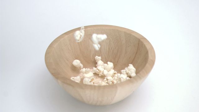 Depicting a few popcorn kernels in slow motion landing into a natural wooden bowl, emphasizing simplicity and focus. Highlights enjoyable casual moments such as movie nights, snacking, or homely activities. Ideal for use in advertising casual snacks, illustrating concepts of relaxation or enjoyment, and food-related blogs seeking an eye-catching image.