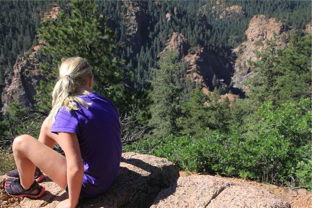Young girl, in casual attire, rests on rocky cliff, enjoying peaceful view of forested canyon. Ideal for depicting outdoor adventures, hiking, exploring nature, travel, and tranquil moments in wilderness.
