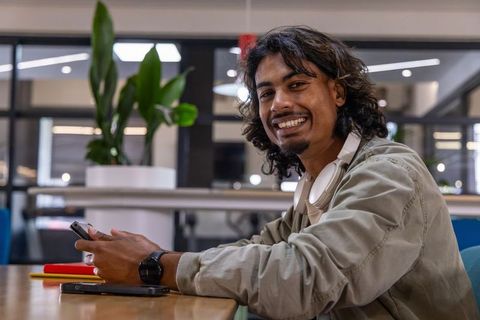 Young asian man smiling while using smartphone in modern coworking space with headphones