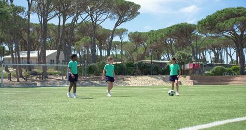 Diverse teenage girls practicing soccer on sunny artificial turf near goal and pine trees