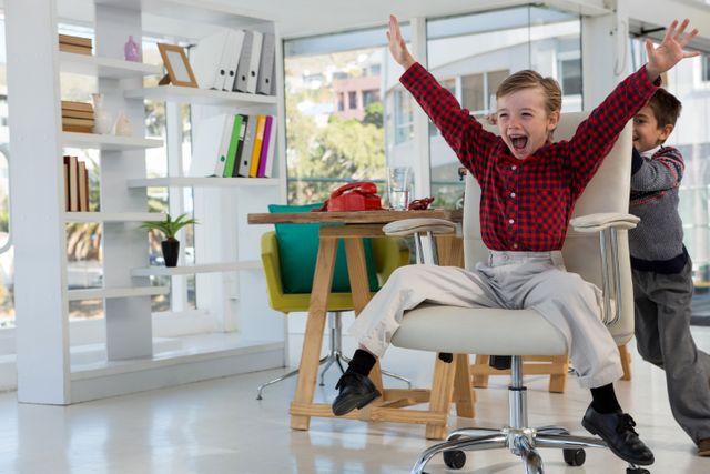Children are seen playing in a modern office environment. One child is pushing another on a swivel chair, both displaying joy and excitement. This image can be used to depict concepts of fun at work, youthful energy, and a casual office atmosphere. Ideal for use in articles about work-life balance, modern office culture, or advertisements for family-friendly workplaces.