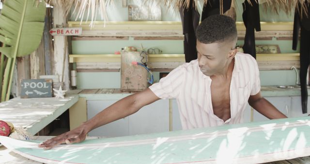 Happy african american man in surf shop on beach preparing surfboard. Lifestyle, business, vacation, summer and leisure, unaltered.