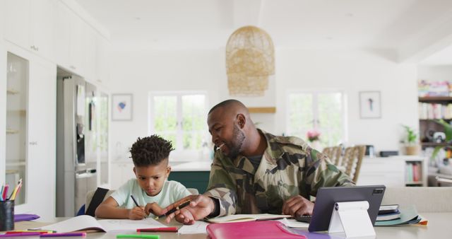 African American army father helping young son with homeschooling at kitchen table. Shows strong family bond, parental support in education, and a modern, well-lit home environment. Ideal for content on family bonding, parental involvement in education, military families, and modern home settings. Can be used in articles, advertisements, and blogs focusing on parenting and education.