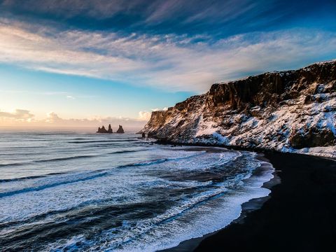 Glowing winter coastline with snow-dusted cliffs, black sand and distant sea stacks at sunrise