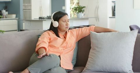 Mature asian woman relaxing on gray sofa wearing white headphones, resting with pillow
