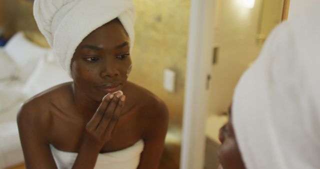 African American woman stands in front of a mirror in her bathroom, wrapping her hair with a white towel while applying skincare products on her face. Perfect for articles and promotions related to skincare, beauty routines, self-care practices, and healthy lifestyle content.