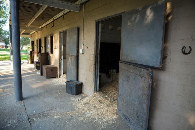This image shows an empty horse stable with open doors, allowing sunlight to stream in. The stable is made of concrete blocks with wooden beams supporting the roof. Hay is scattered on the ground, and a horseshoe is hung on the wall. This image can be used for themes related to rural life, agriculture, equestrian activities, or farm management. It is ideal for illustrating articles, blogs, or advertisements about horse care, farming, or countryside living.