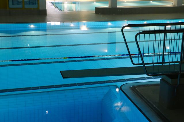 Indoor swimming pool with clear blue water, lane markers for laps, and a diving board. Pool area is well-lit with modern lighting fixtures. Ideal for use in advertising fitness centers, aquatic training facilities, or leisure activities.