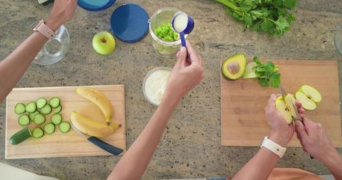 Overhead hands preparing green smoothie ingredients apple avocado cucumber banana yogurt