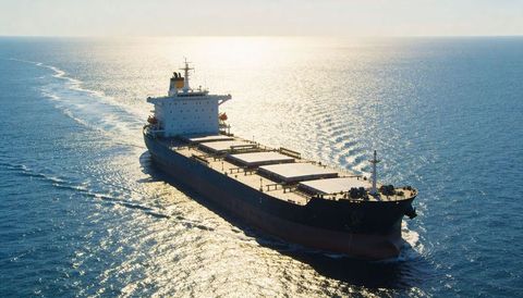 Bulk carrier cutting through sunlit open sea leaving wake, exposing hatch covers and deck