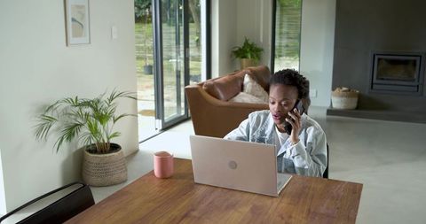 African american woman working from home on laptop while talking on phone in sunlit open-plan home