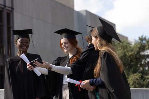 Diverse female graduates celebrating and taking selfie on university campus plaza