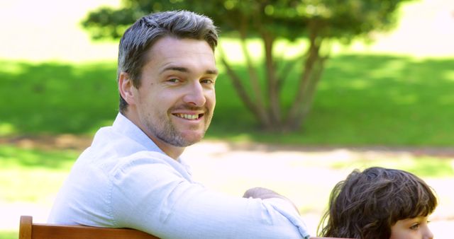 Man is smiling while sitting on a bench in a park. He is with a child that is partially visible. Sunlight illuminates the scene, enhancing the green natural background with trees and grass. Ideal for use in family-focused articles, parenting blogs, advertisements for outdoor activities, or promotional materials for parks and recreation areas.