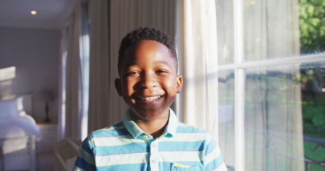 Young African American boy happily smiling near a sunlit window, dressed in a light blue and white striped shirt. Perfect for use in projects related to home life, children's activities, advertising, or educational materials featuring happiness and the warm feeling of home.