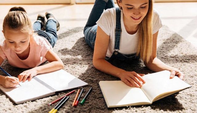 Sisters drawing and reading together on cozy sunlit carpet at home, creative family moment