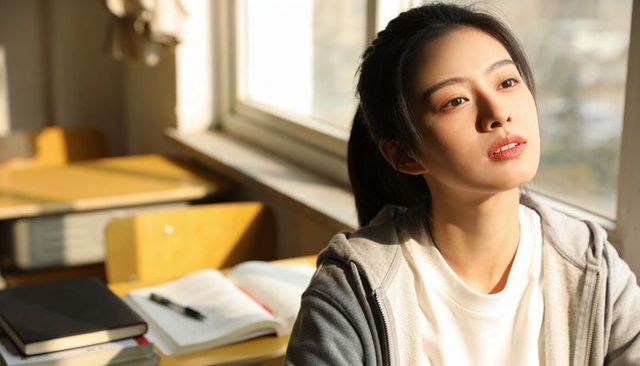 Young student gazing out sunlit classroom window during study break, notebook on desk
