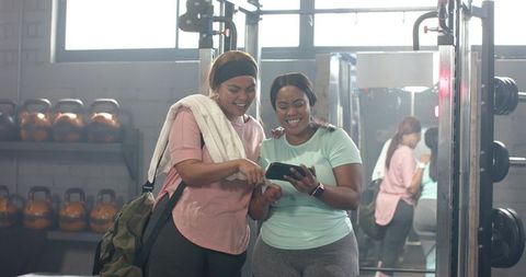African american women sharing workout plan on smartphone at gym, smiling and encouraging