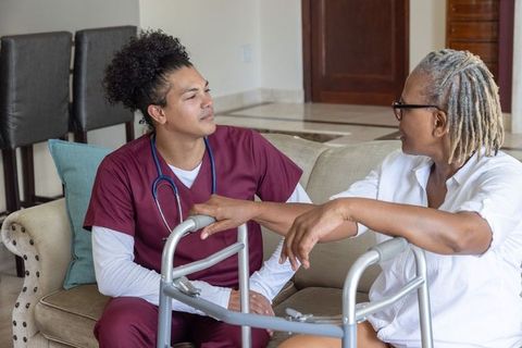 Home caregiver nurse talking with african american senior using walker on living room sofa