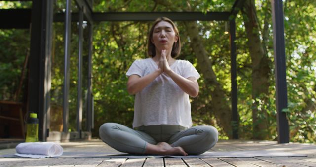 Woman is in a cross-legged yoga position on a wooden deck surrounded by lush, green forest. She is meditating with eyes closed and hands in a prayer position. Green water bottle and white towel are nearby. Suitable for wellness blogs, meditation apps, fitness magazines, and stress-relief advertisements.