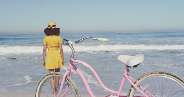 Woman in yellow dress facing the ocean, enjoying the scenic view, with a vintage pink bicycle in the foreground. Ideal for travel, summer, and leisure lifestyle themes. Could be used in advertisements, blog posts, or social media content promoting beach vacations, relaxation, and outdoor activities.