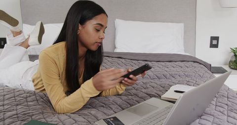 Young woman lying on bed multitasking with smartphone and laptop for remote work and study