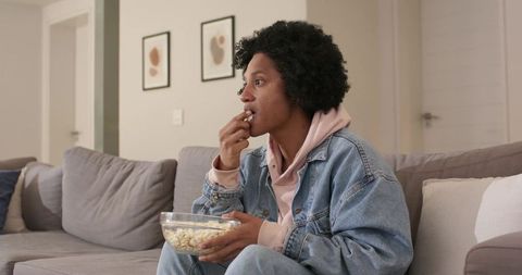 African-american man sitting on sofa eating popcorn while watching tv in cozy living room