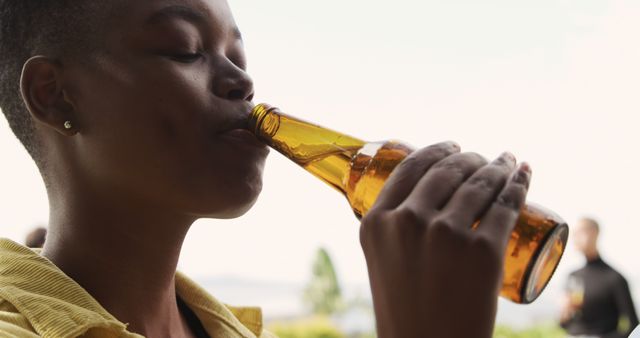 Young person drinking beer from bottle while spending time outdoors on a sunny day. Suitable for campaigns on leisure, beverage promotion, lifestyle journalism, relaxation concepts. Effective for social media posts, advertisements, and websites that focus on casual drinking, outdoor activities, and personal enjoyment.