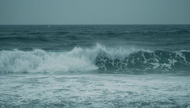 Powerful teal ocean wave crashing onto shore with white foam and overcast sky