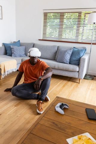 Young man wearing vr headset sitting on hardwood floor near couch with game controller
