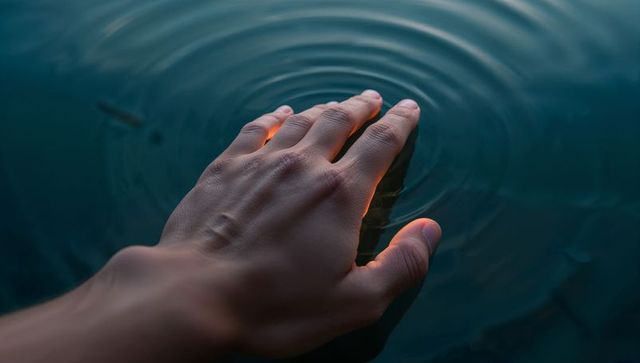 Hand touching calm pond creating concentric ripples and subtle warm rim glow at dawn